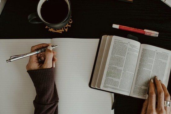 A woman's left hand sits on top of an open notebook with a pen, ready to write; her right hand rests on an open Bible