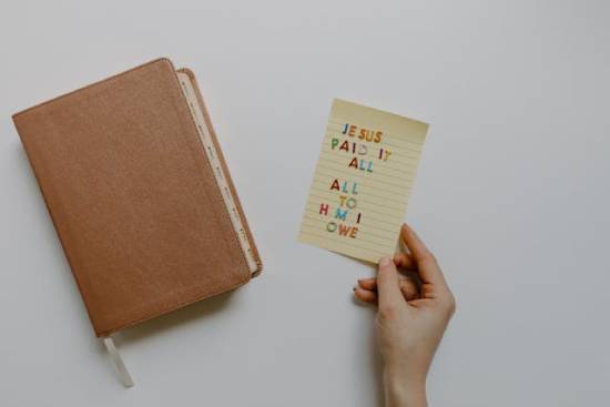 A hand holding a paper beside a closed Bible that reads "Jesus paid it all, All to Him I owe." Someone holds a note that says, "JESUS PAID IT ALL, ALL TO HIM I OWE" next to their closed Bible. Jesus fulfilled all the requirements for our salvation and transformation.