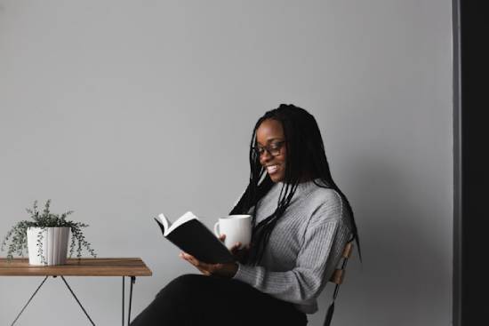 A young woman happily sits and reads a book while drinking a hot beverage.