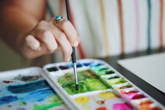 A woman's hand dips a small paintbrush into a paint palette.