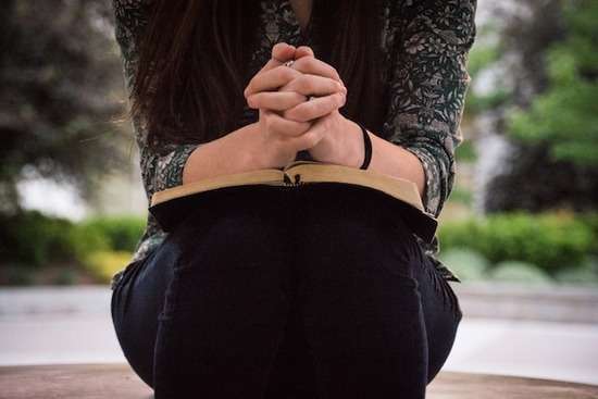 Woman sitting while praying with folded hands on an open Bible as we learn about various physical postures during prayer