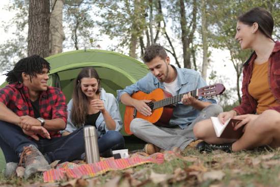 Group of four young people camping with one playing a guitar Four friends hanging out at a campsite while one of them plays a guitar. This illustrates the sense of fellowship and community Adventists may experience at camp meetings.