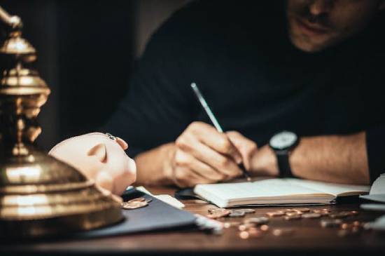 a man sitting at a desk with a piggy bank on it, in a darkened room lit by a desk lamp, recording in his ledger.