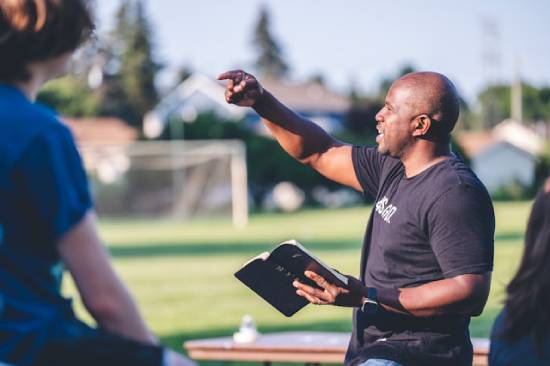 A man holding an open Bible and gesturing with his right hand, showing that he is sharing a biblical lesson or principle to people in front of him.