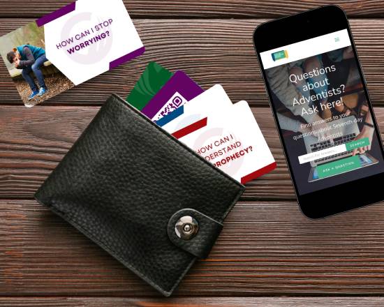 A wallet with several Ask An Adventist Friend cards fanned out sits on a table with a smartphone on the right and an Ask An Adventist Friend card that says 