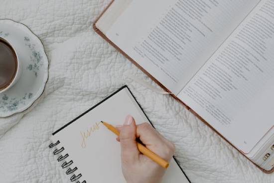 A woman sits on a bed with her morning coffee, an open Bible, and a notebook. She writes "Jesus" at the top of a page, taking note of what she's learning about Him as she studies the Word.
