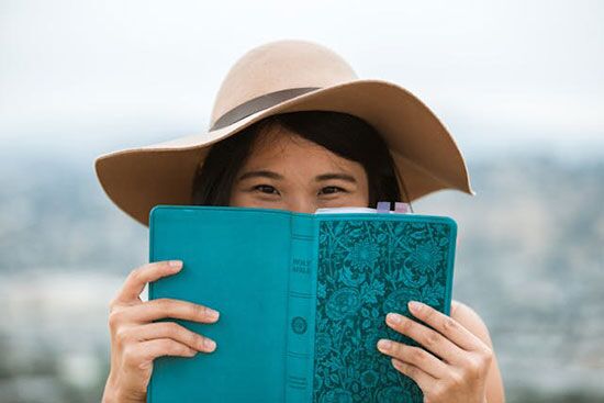 Woman holding a Bible in front of her face a young woman with a hat on smiles with her eyes, an open Bible covering the bottom half of her face.