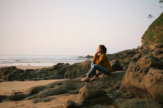 A woman meditating on her thoughts near the sea A barefoot young woman sits on the rocks at the beach and contemplatively gazes into the sunset