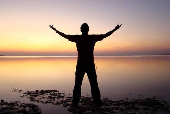 A Man Looking in Heaven Raising Arms A silhouette of a man standing in front of a sunset over a lake with his arms reaching up. This is a common image to illustrate spiritual health.