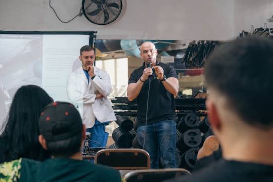 A man speaks with a microphone to a small group; a doctor stands behind him.