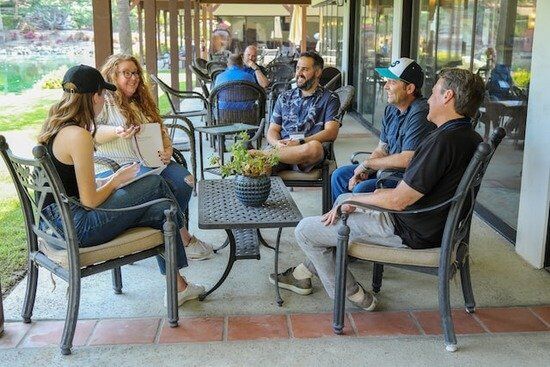 a small group of friends sits around a table outdoors, talking and laughing; two other men sit talking to one another in the background