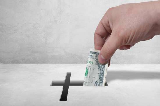 A hand places a folded $1 bill into a cross-shaped slot, signifying that a small portion of our income should be dedicated to God, as He provided the ultimate sacrifice for us.