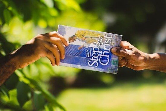 two hands exchanging a Steps to Christ book by Ellen White A hand giving Ellen White's book Steps to Christ to someone
