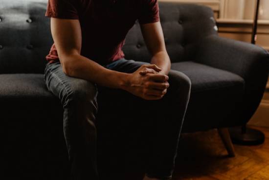 Pensive, struggling man sitting on a couch A man sits on a couch with his hands clasped together, illustrating the pressure of personal struggles.