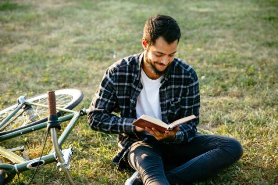 Man sitting on the grass beside his bicycle reading the Bible A man sits in a green field next to a bike while reading his Bible—a reminder that the Bible is our ultimate source of truth.