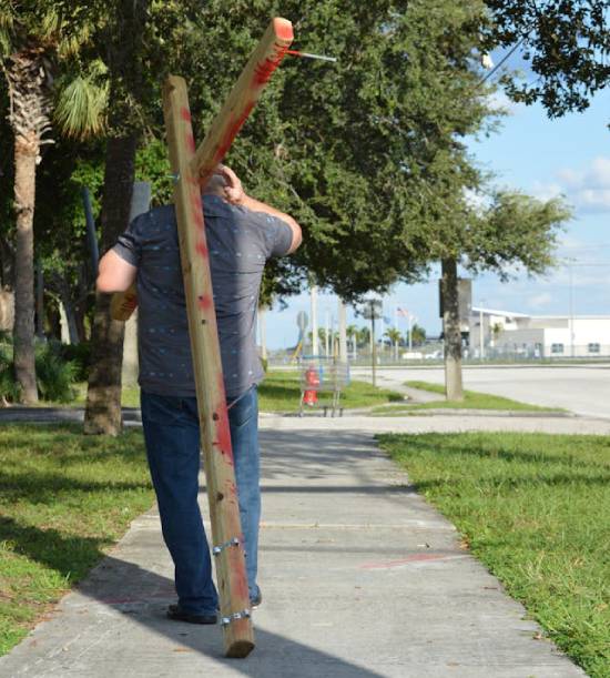 Back view of a man walking on a sidewalk while carrying a large cross A man carries a cross down a modern-day sidewalk, making us wonder what it really means to pick up your cross like Jesus.
