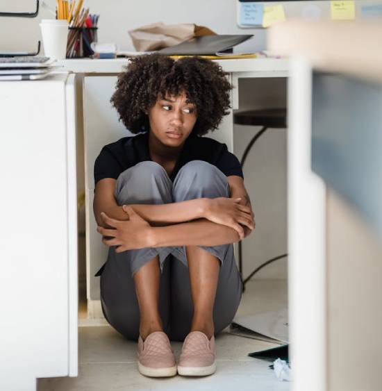 Sad woman sitting on the floor beside her desk A sat woman sits under a desk with her arms wrapped around her knees, highlighting what Jesus didn't meant by taking up our crosses.