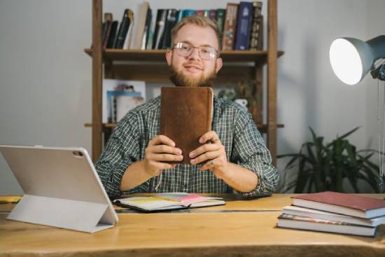 A happy man holding his Bible that is full of encouragement A young man happily sits at a desk holding up his Bible.