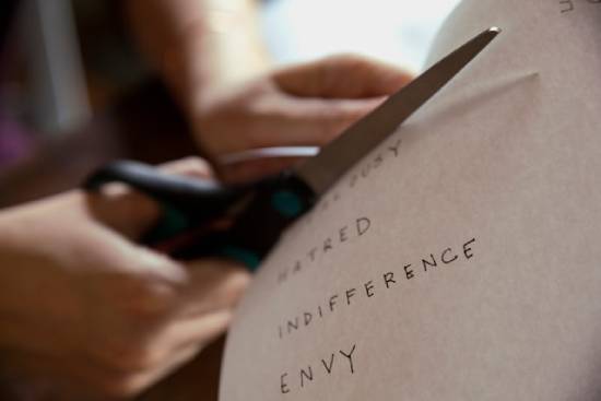 Words like jealousy, hatred, indifference, and envy written on a paper being cut A person cuts out words of negative character traits (e.g. hatred, indifference, envy) from a piece of paper.