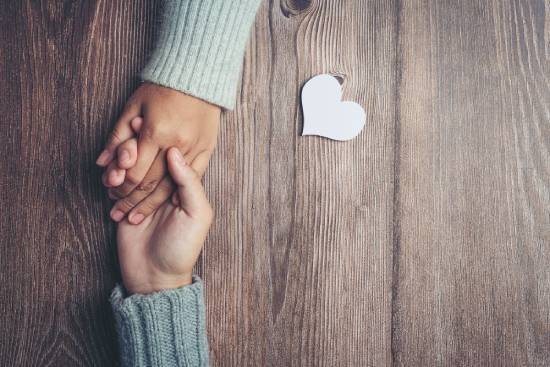 Two people holding hands in an encouraging way Two hands interlock on a table with a paper cutout heart nearby, indicating love and care between them.