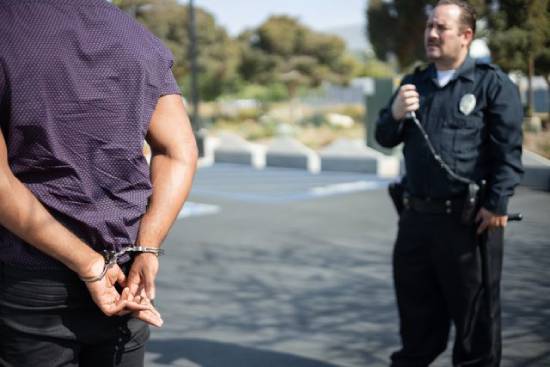 A man in handcuffs while a police officer talks on his radio A man in the foreground stands in handcuffs while a police officer in the background looks on.