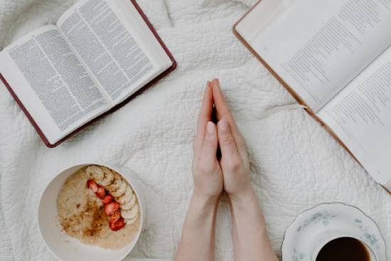 A woman's hands are folded while surrounded by Bibles, a bowl of oatmeal, and a cup of tea. This illustrates how faith is a lifestyle that's shaped by God's Word and the Holy Spirit.