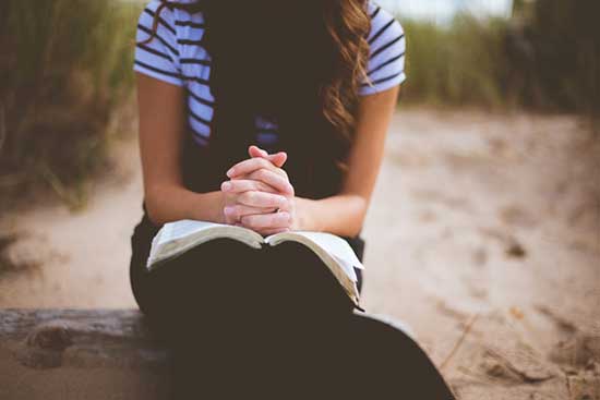 A modestly dressed woman with her hands folded in prayer over her open Bible.
