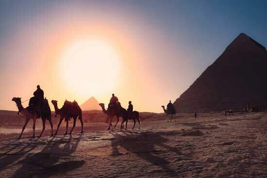 Travelers passing by the Egyptian pyramids while riding on camels and a horse.