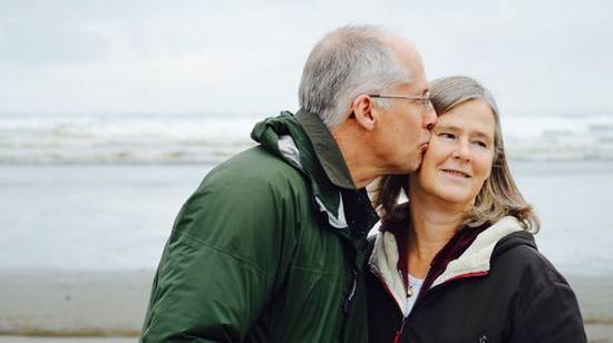 A gray-haired man kissing his wife on a beach and enjoying a quality life because of good health habits and trust in God.