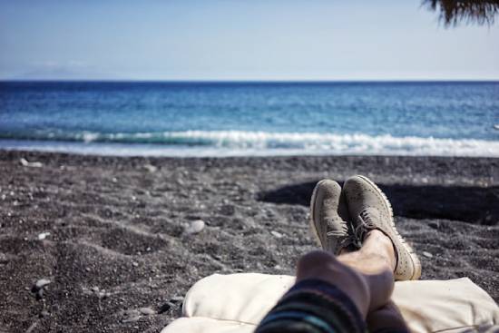 A man's laid-out legs can be seen on a lounge chair on the beach in front of an ocean.