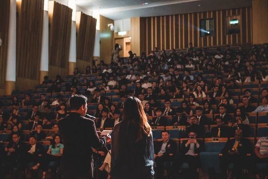 A man and woman speak in front of an auditorium full of people.
