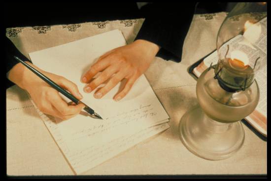 A woman sits at a table writing by a burning oil lamp.