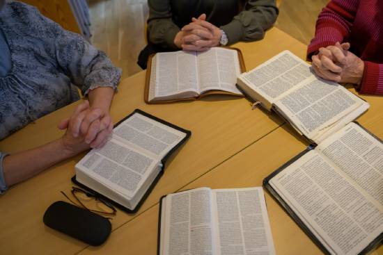 Three people sit around a table with their Bibles open and their hands clasped in prayer; a pair of glasses rests near one of the open Bibles