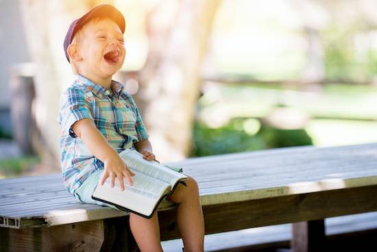Young boy sitting outdoors holding an open Bible and laughing A little boy holding a Bible and smiling