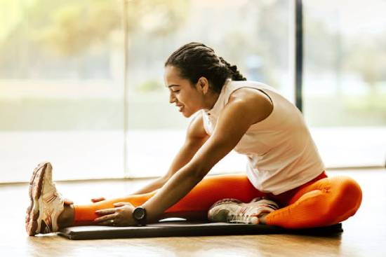 A woman stretches her leg while sitting on the floor, demonstrating fitness as a necessary part of our health.