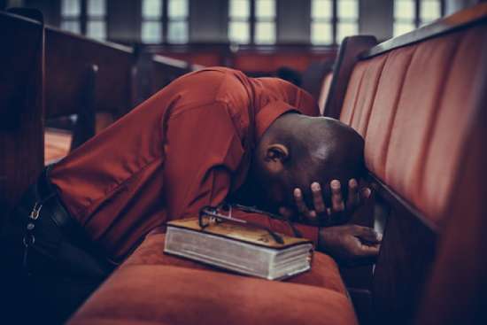 A man kneels and prays in a church pew with a Bible beside him, illustrating when we realize we need spiritual healing.