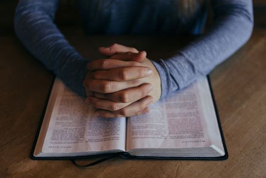Bible Study Person praying with folded hands on a Bible, as we prayerfully consider Biblical principles to guide our decision on tattoos.