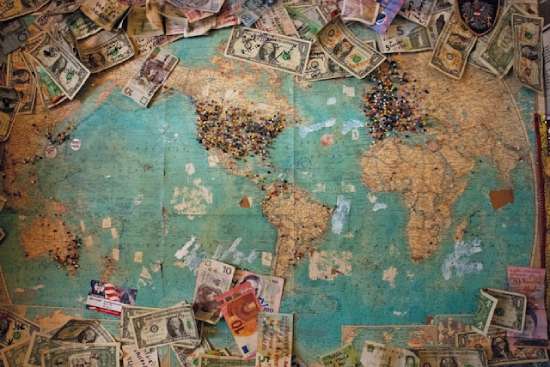 Distribution of money A map spread across the table, pins are placed in various locations, congregating across North America, Western Europe, and Australia. Scattered bills of all denominations border the map.