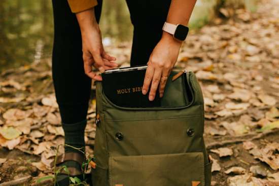 Woman taking her bible out of her backpack A green backpack sits at someone's feet. The person is leaning over to pull a Bible out of the backpack, highlighting the importance of searching the Word of God for answers, not the world.