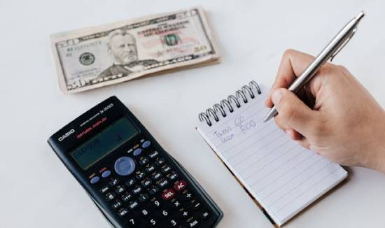 Calculator, money, and budget list a calculator and a small stack of dollar bills sit on a white table, with a person's hand writing in a small open notebook