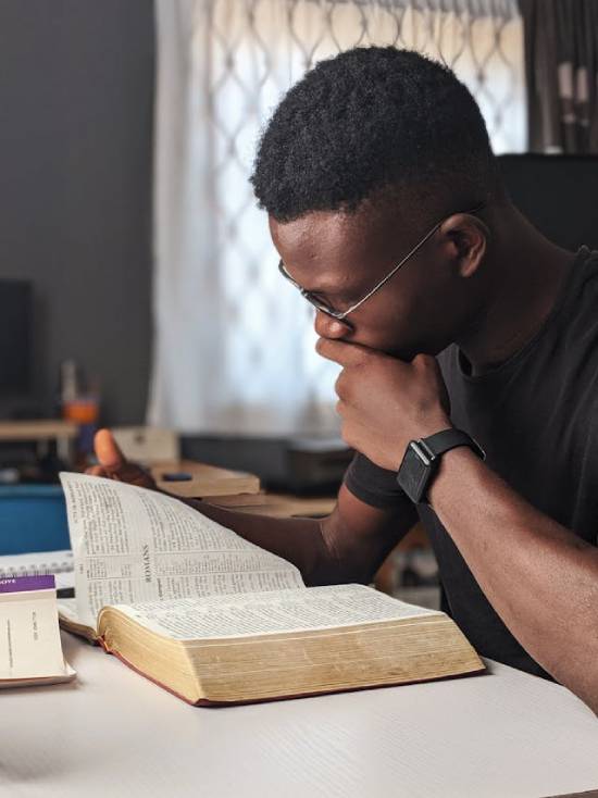 Man Reading a Bible a black man wearing glasses and sitting at a table, with his hand on his chin studying an open Bible, in deep thought