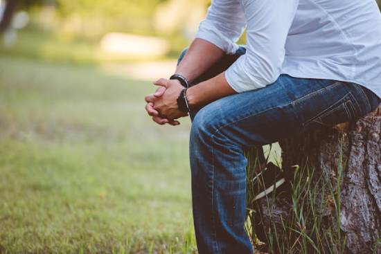 Man praying outdoors while sitting on a stump. A man sits on a log outside with his hands folded in prayer.