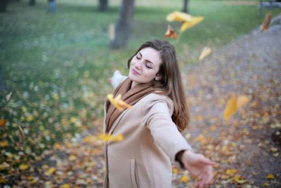A woman stands, with arms outstretched on an outdoor walkway. Yellow autumn leaves float around her.