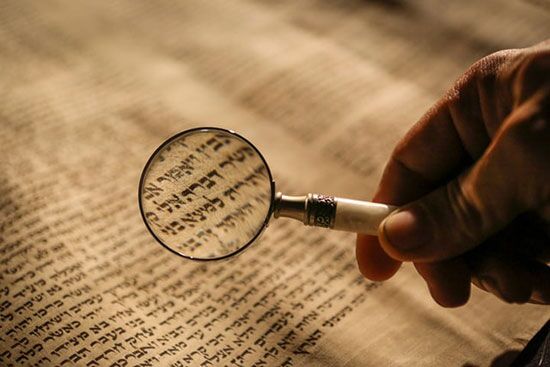 A hand holding a magnifying glass to Hebrew scrolls