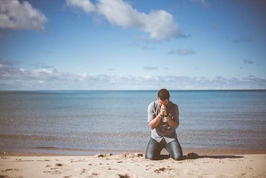 Kneeling Prayer Position Man kneeling down at seashore praying as we learn about the various physical postures during prayer as mentioned in the Bible
