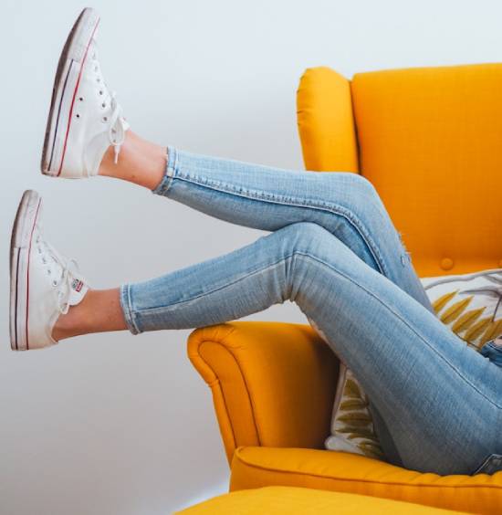A woman kicks her legs kicked over the side of an orange chair while wearing skinny jeans and white shoes, which represents how clothing should enhance, and not harm, our health.