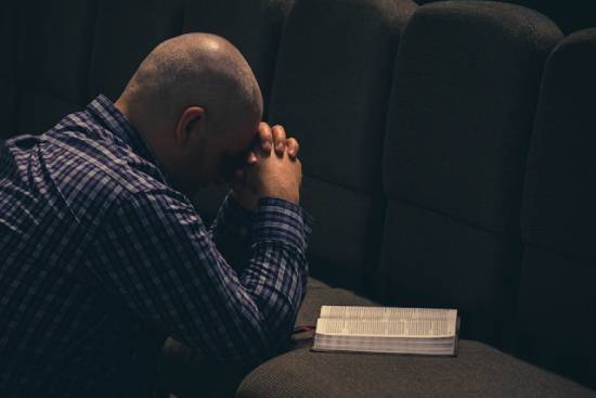 Man kneeling and praying beside an open Bible Man bowing head and clasping hands in prayer before an open Bible