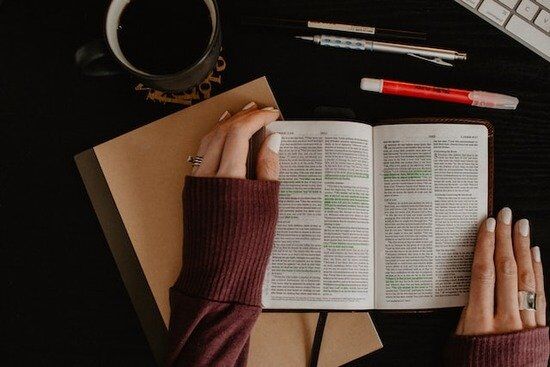 Studying Bible Hands holding a Bible open with pens and other study materials beside