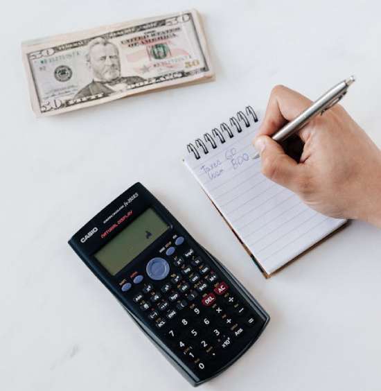 Calculator and Money on a Table A person writes in a notebook beside a calculator and a stack of money as they evaluate their finances.