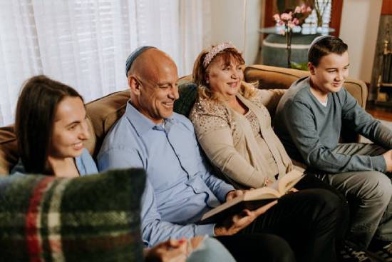 A Jewish family sits on a couch to welcome the Sabbath as the father opens the Bible to read from it.